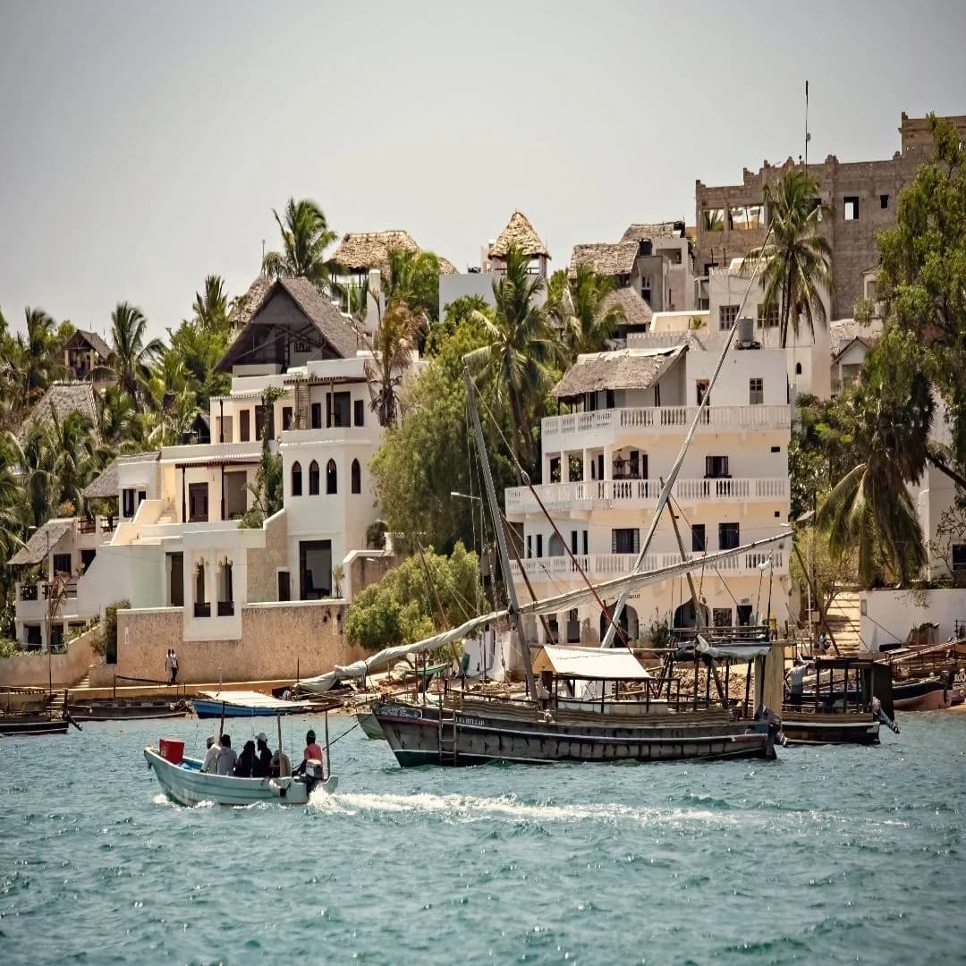 Traditional dhow boats sailing along the historic coastline of Lamu Island with coral-stone buildings and palm trees at sunset